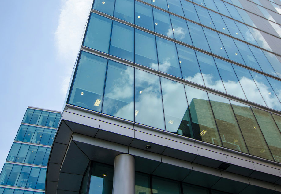 A close-up view of a modern glass office building with reflective windows and a visible steel support column, set against a clear sky.