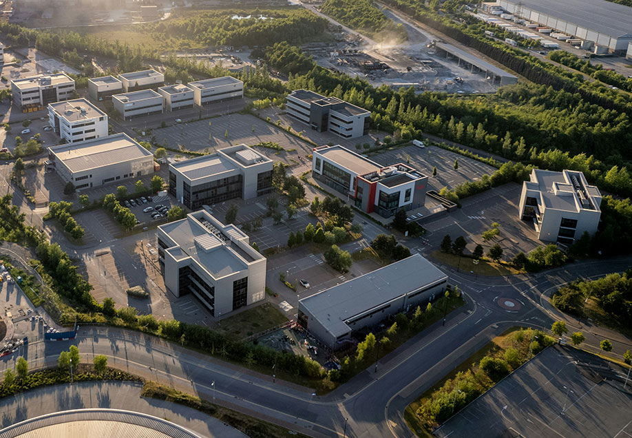 Aerial view of a business park with modern office buildings, wide roads, and green landscaping, surrounded by forested areas and industrial development.